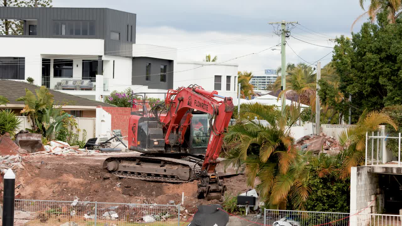A red excavator removes palm trees and debris at a residential construction site in Gold Coast, Australia. Daylight, wide shot, steady camera