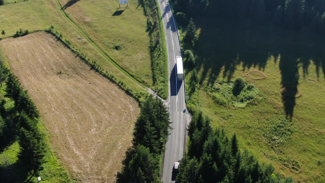 An aerial shot shows a road running through a forest with a large truck on it next to a parking area with several cars parked