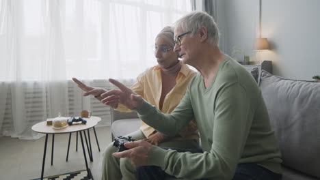 mujer árabe de mediana edad explicando a un anciano cómo usar el controlador de juego mientras se sientan juntos en el sofá en casa 1
