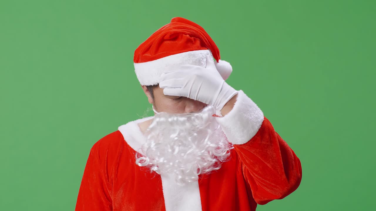 녹색 스크린 앞에 서 있는 아시아 산타클로스가 두통을 고 있습니다.