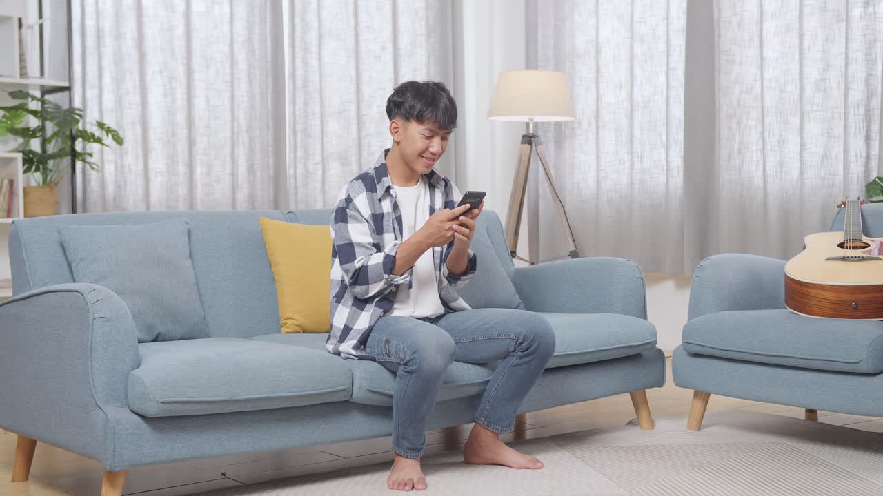 아시안 십대 소년의 몸이 가득 차 있고 집에서 거실에서 소파에 앉아 스마트폰을 즐기고 있습니다.