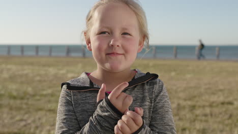 close up portrait of playful little blonde girl smiling happy enjoying sunny seaside park looking at camera shy