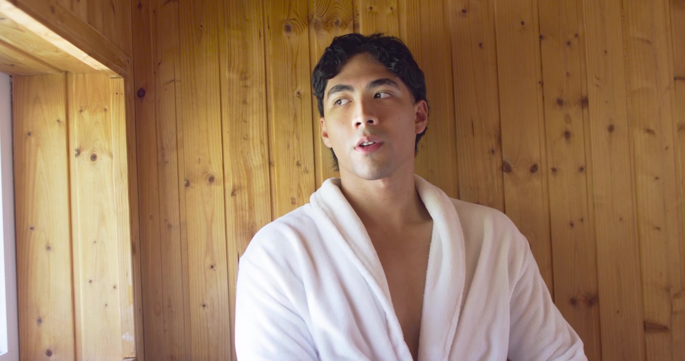Adult male sitting in white bathrobe in wood-panel room glancing from window, turning toward camera