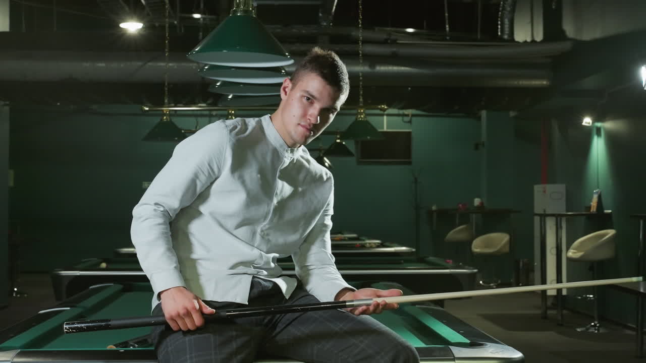 игрок в белой рубашке и серых брюках сидит на зеленом бильярдном столе с легкой улыбкой, держа в руках палку. верхние лампы, современная бильярдная зала с несколькими бильярдными столами на заднем плане