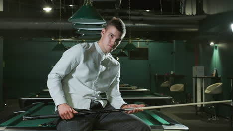 player in white shirt and grey trousers sits on green pool table with light smile, holding cue stick. overhead lamps , modern billiard hall setting with multiple pool tables in background