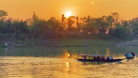 two passenger boats are passing through the surma river with people loaded on them with a beautiful golden sunset in the background