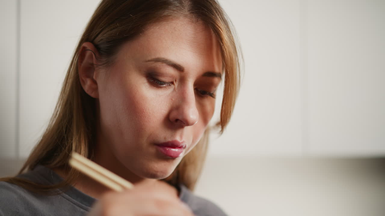 Close up of lady eating meat in modern kitchen, drops meat into plate then picks another piece to eat, calm facial expression, soft blur white cabinetry in background