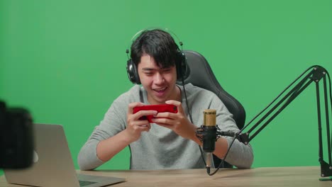 a smiling asian man gamer with headphone and computer looking at camera while using mobile phone playing game on the green screen background