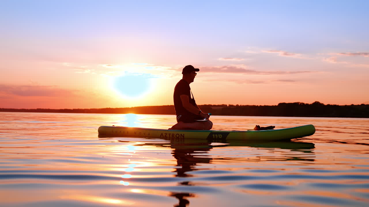 Man sits on his knees on sup board holding an oar in hands. Sportsman floats by the water looking at beautiful sunset.