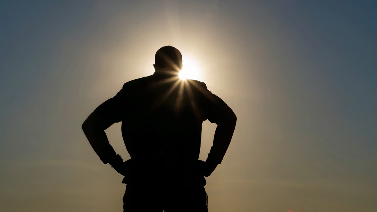 Uomo in piedi che osserva l'alba da un punto panoramico, il sole che si sposta dietro la testa creando un alone luminoso, in giacca.