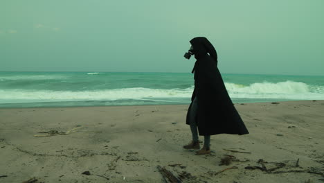 a man in a cloak and gas mask walks on a radiation poisoned beach