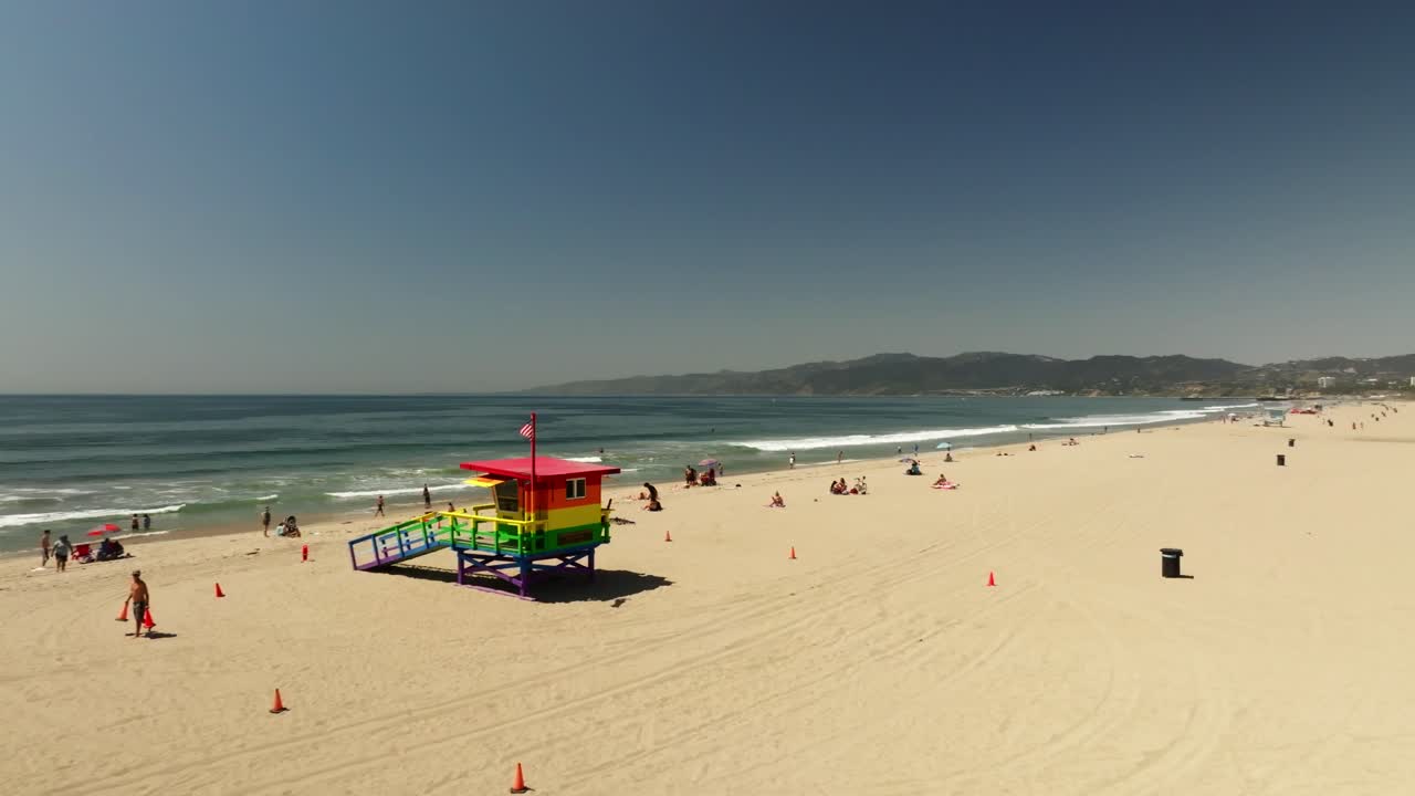 Panoramic views of Venice Beach featuring a vibrant rainbow lifeguard tower, the ocean, and people enjoying the sunny day.