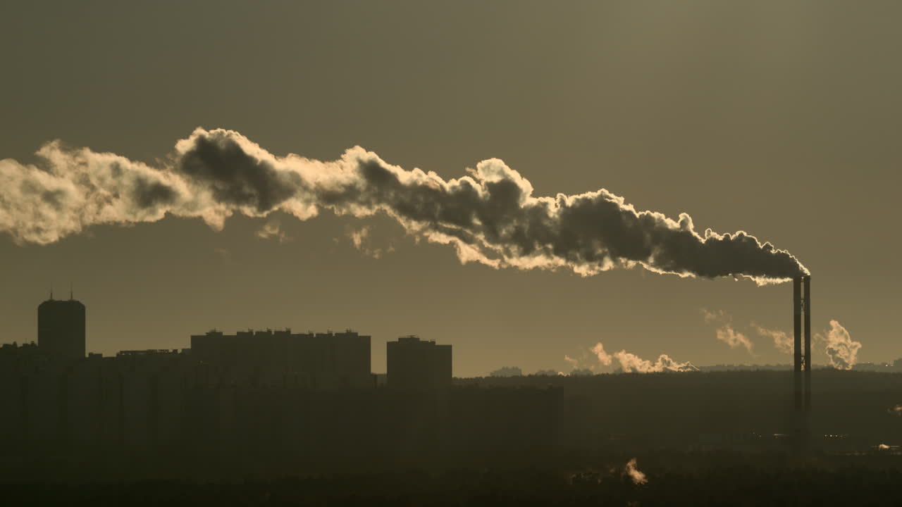 Chimenea industrial alta liberando una larga y densa columna de humo sobre el horizonte residencial con una cálida luz de fondo dorada. Escena urbana con teleobjetivo, neblina, emisiones de infraestructura de calefacción urbana y sensación de contaminación atmosférica