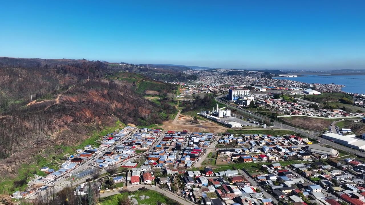 Lento paljastaa rannikkosatamakaupungin tiheine asuinalueineen, teollisuussiiloineen ja palaneine rinteineen, jotka ulottuvat lahdelle ja kaukaiselle satamalle. Lirquén, Penco, Chile