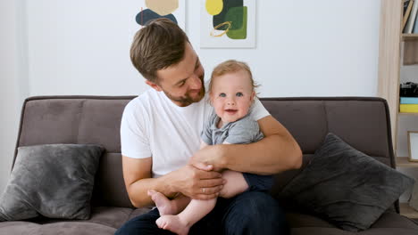 hombre guapo feliz sentado en el sofá en la sala de estar mirando a la cámara durante una videollamada mientras abraza a su lindo niño 1