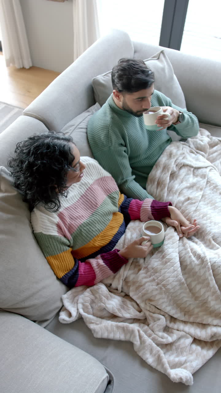 Video verticale di coppia multietnica seduta sul divano e che beve caffè a casa, al rallentatore.
