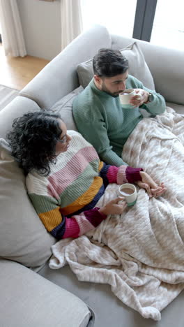 vertical video of biracial couple sitting on sofa and drinking coffee at home, slow motion
