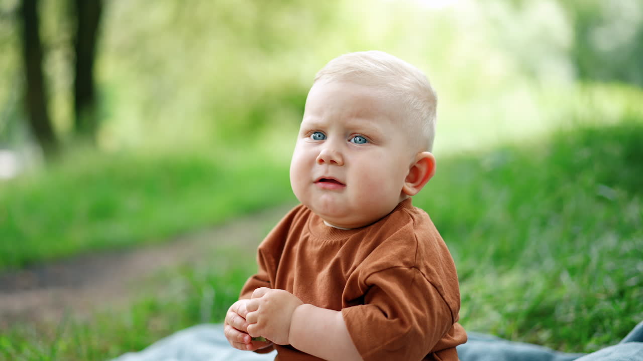 Blue-eyed blond toddler sitting outdoors. Kid is holding a tree leaf in his hands and showing it. Blurred backdrop.