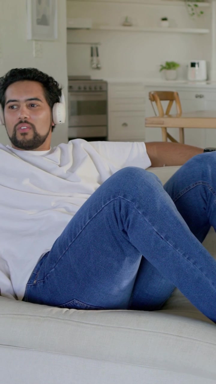 Video verticale: Uomo sdraiato con una maglietta bianca e jeans che reagisce all'audio nelle cuffie a casa.
