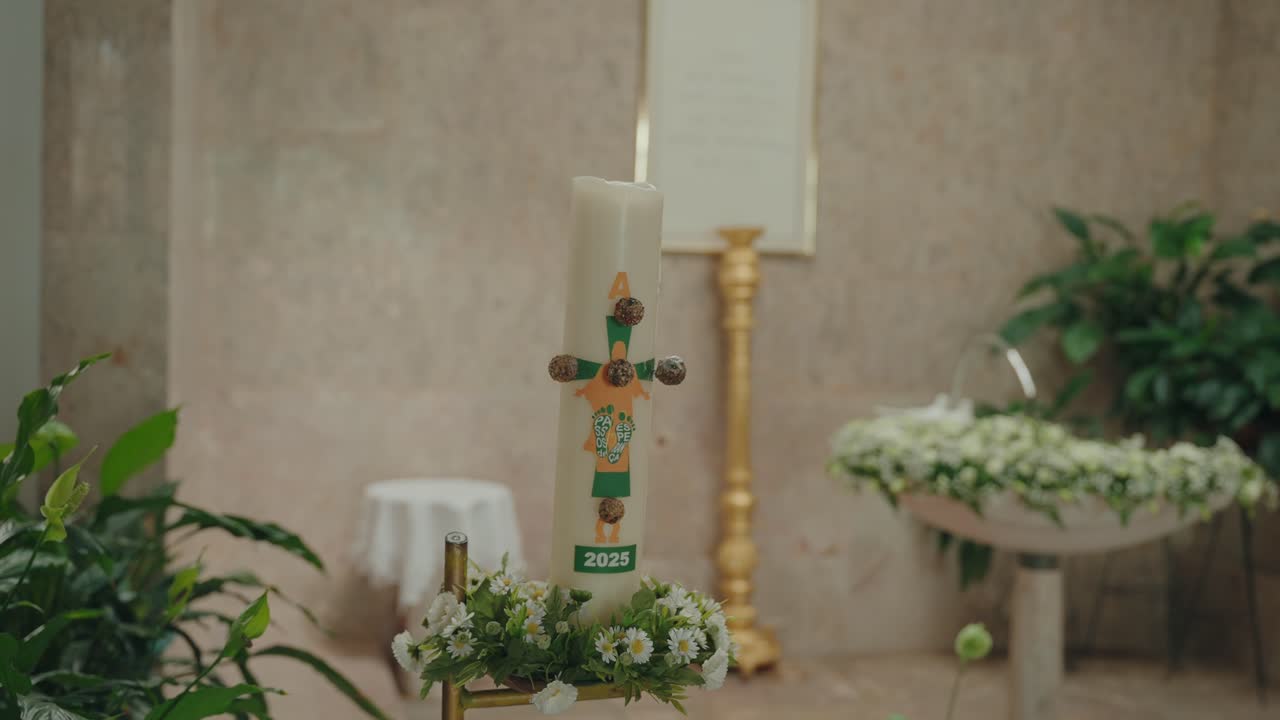 Primer plano de un cirio pascual con un arreglo floral cerca de la pila bautismal en una iglesia