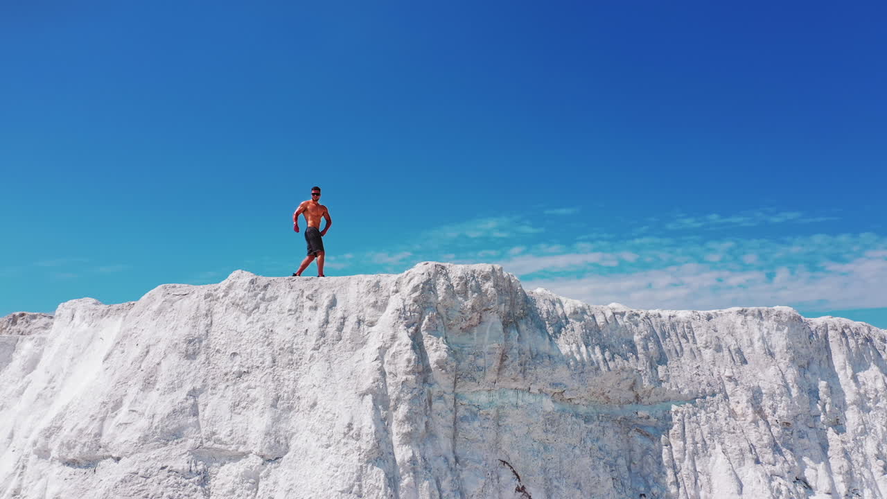 Muscular sportsman under blue sky. Strong athlete in black sunglasses posing on camera on the top of white mountain. Camera moves forward. Drone view.