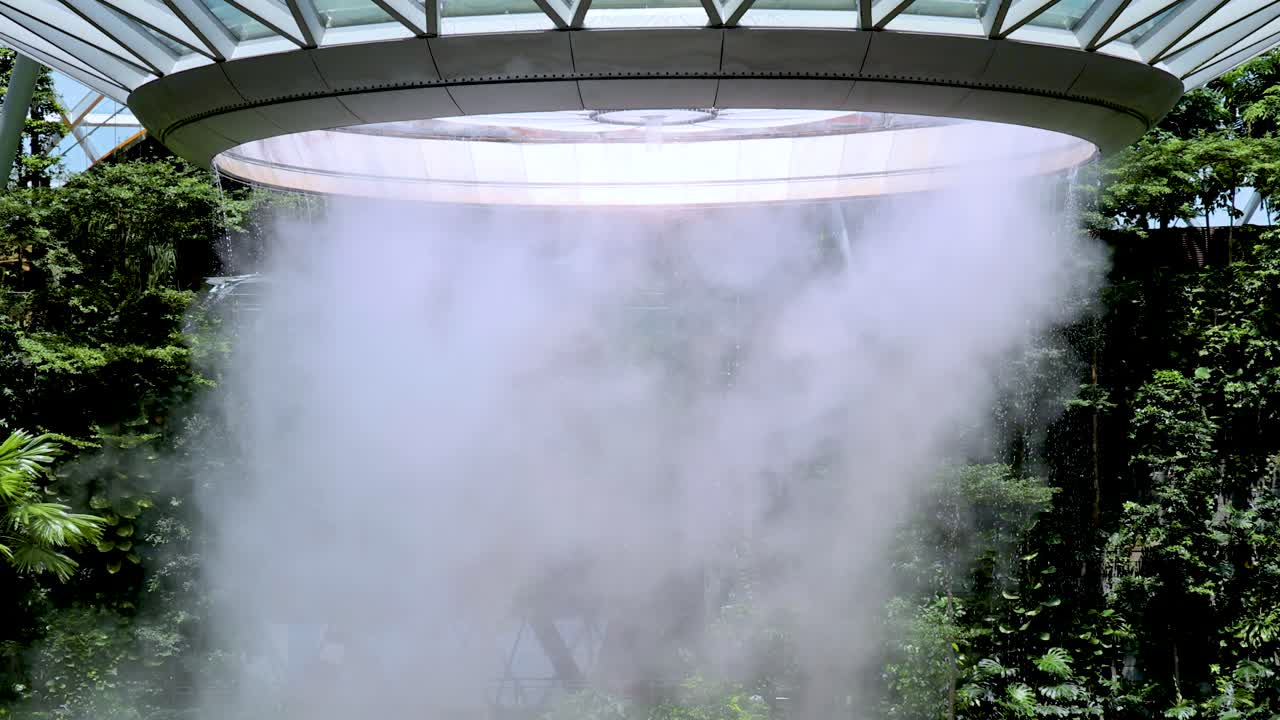 A dramatic indoor waterfall pours from a circular skylight into a verdant, glass-roofed conservatory, surrounded by dense tropical foliage and natural daylight