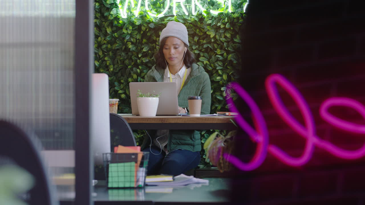 giovane donna d'affari afroamericana che usa un computer portatile, naviga online, scrive messaggi, beve caffè, ascolta musica, controlla lo smartphone in un ufficio alla moda