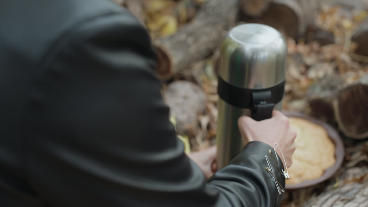 woman in leather jacket pouring thermos beside log stack, closeup of hands unscrewing cap and gripping stainless bottle, metal bracelet with safety pin details visible, pie on wooden plate resting