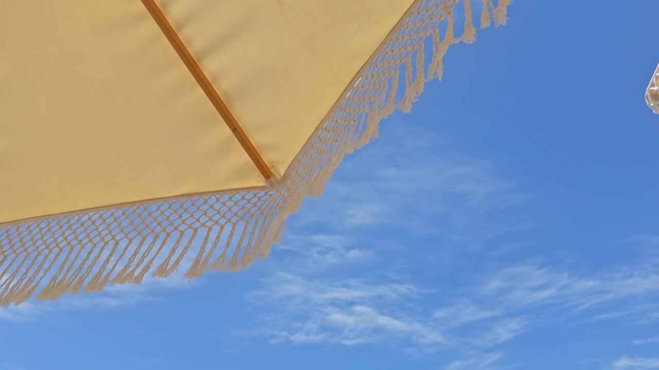 Splendida ripresa 4K dal basso di ombrelloni in macramè bianchi con frange in un resort di lusso sulla spiaggia. Presenta una fila organizzata di lettini, palme tropicali e un cielo azzurro limpido