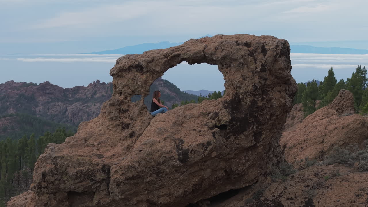 Aerial footage capturing a woman framed by the Window of Roque Nublo (Gañifa), with Mount Teide completing the scene. Gran Canaria, Canary Islands.