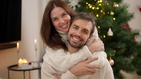 Portrait-of-a-Happy-guy-and-a-brunette-girl-in-a-White-sweater-who-hugs-her-boyfriend-from-behind-and-smiles-in-a-cozy-Christmas-decorated-room-with-a-Christmas-tree