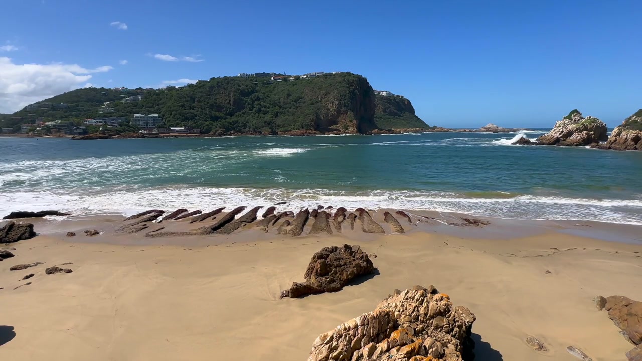 Ondas quebram contra a costa rochosa na Praia Coffin sob um céu azul claro em Knysna, África do Sul.