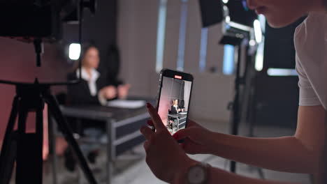 female hands with red manicure hold the phone taking video of a woman sitting and talking at desk. footage of a blog in modern studio.