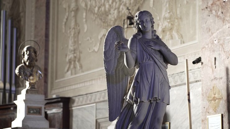 Statua di angelo in marmo illuminata da una luce laterale soffusa all'interno di un'antica chiesa cattolica italiana con pareti scolpite e un busto sullo sfondo