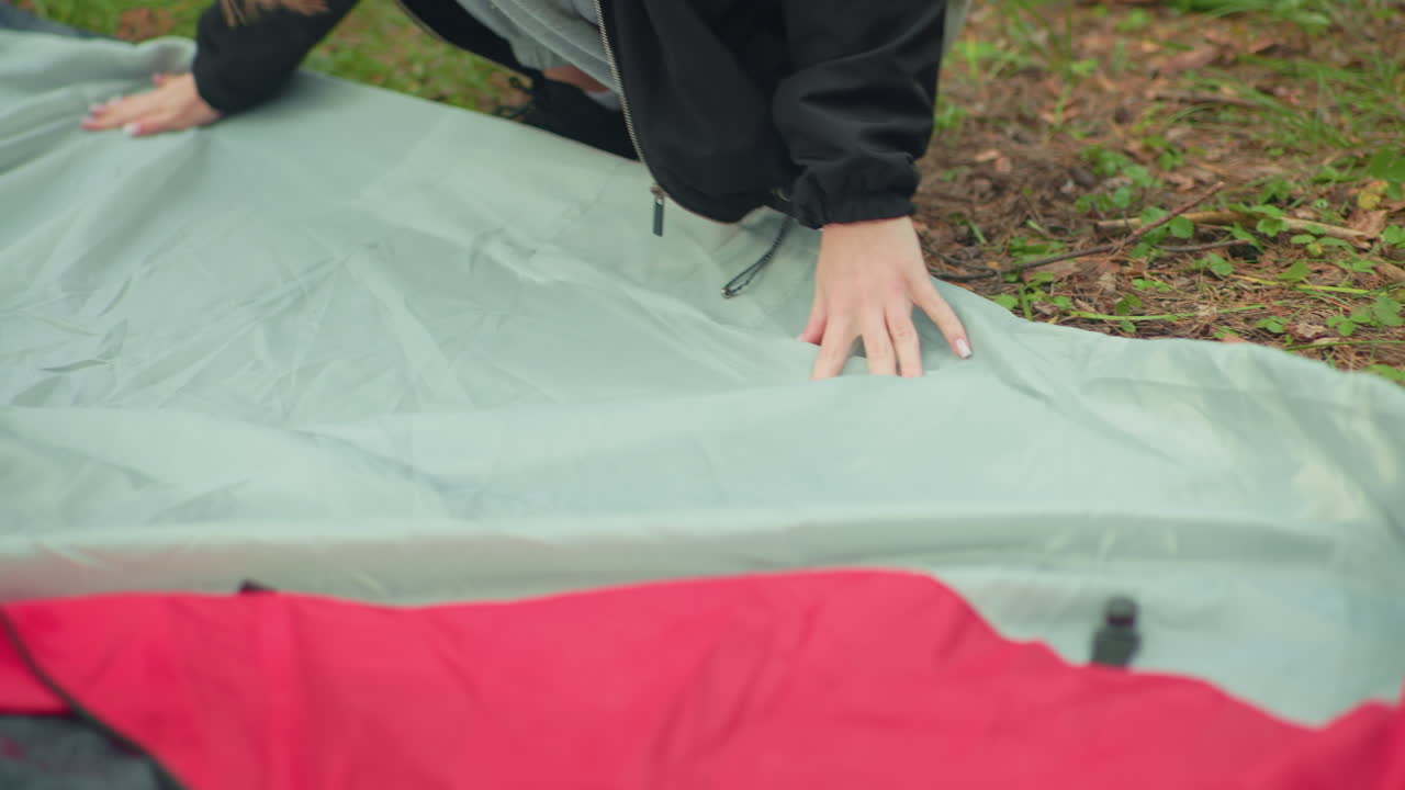 Close up of lady squatting outdoors wearing beanie and black jacket while adjusting tent fabric on grass, with slight background activity of someone spraying tent bag during camping setup process