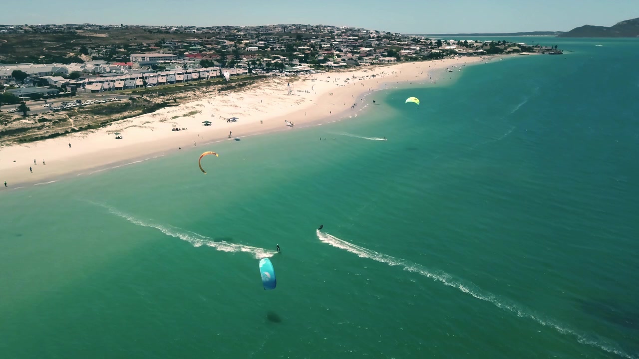 Güney Afrika'daki Langebaan plajında, mükemmel güneşli tatil havasında uçurtma sörfü yapanların yüksek irtifa drone ile çekilmiş geniş manzarası. Su sporları aktiviteleri, berrak okyanus ve mavi gökyüzü ile plaj tatilleri.