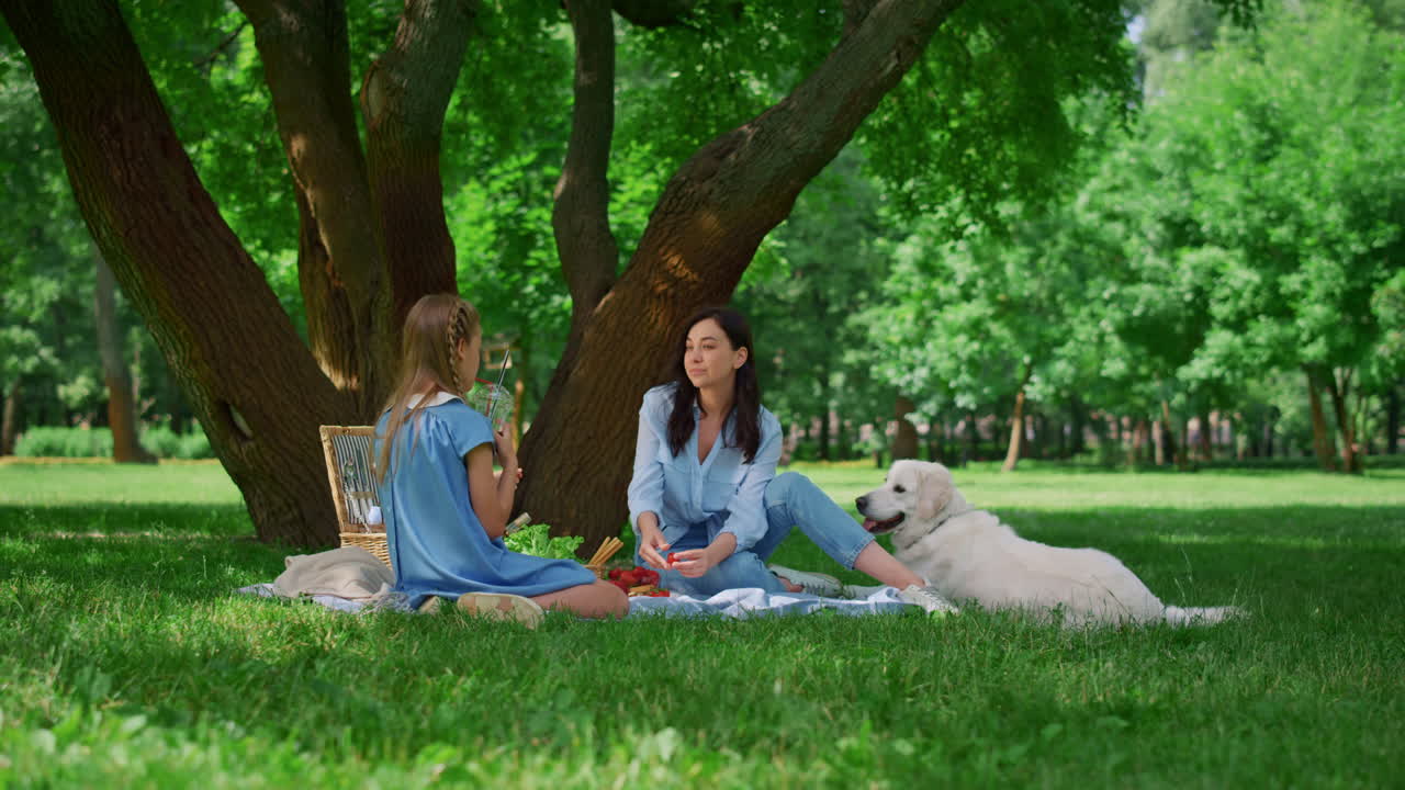 Glad mor hviler med datter på picnic. Familie med hund slapper af udendørs.