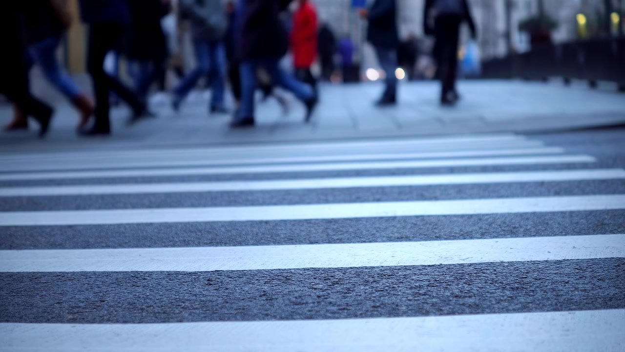 Pedoni che attraversano le strisce pedonali, muovendosi attraverso la strada, in un ambiente urbano probabilmente un centro città o una zona trafficata, con linee pedonali, traffico pedonale, con pavimentazione in cemento e texture di strada asfaltata.
