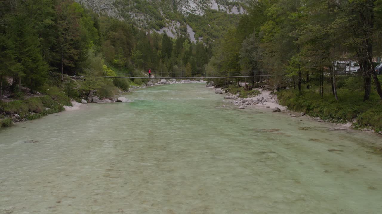 Young woman walk across suspension bridge above a calm river at Soca valley, aerial