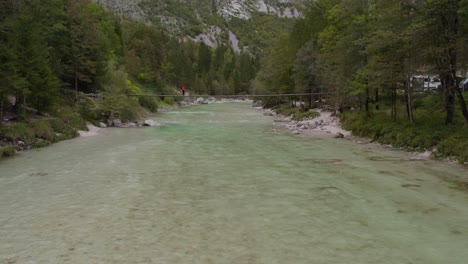 mujer joven camina a través del puente colgante sobre un río tranquilo en el valle de soca, aérea