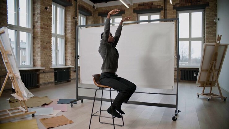 Seated model beginning arm raise, modeling in studio, wearing gray sweater on stool beside backdrop