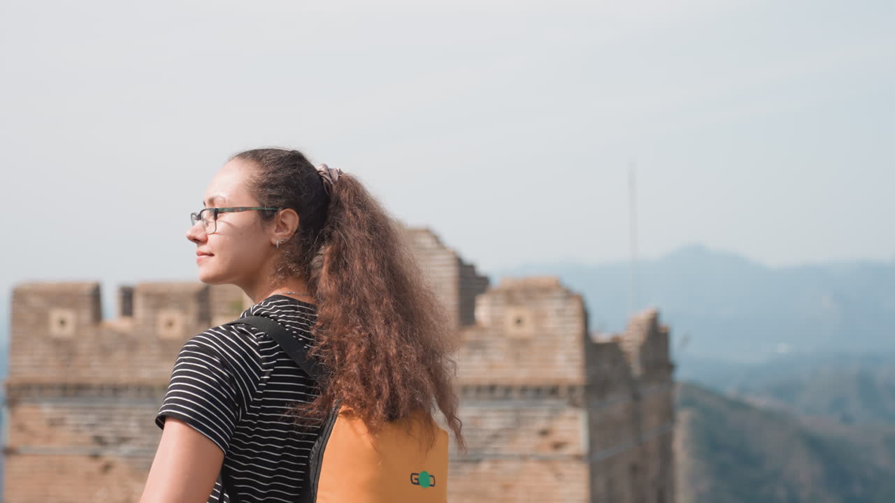Individuo con zaino luminoso osserva una fortezza e montagne distanti. Escursionista donna con zaino giallo vivace si ferma per osservare montagne aspre e una roccaforte lontana nella tranquilla campagna.