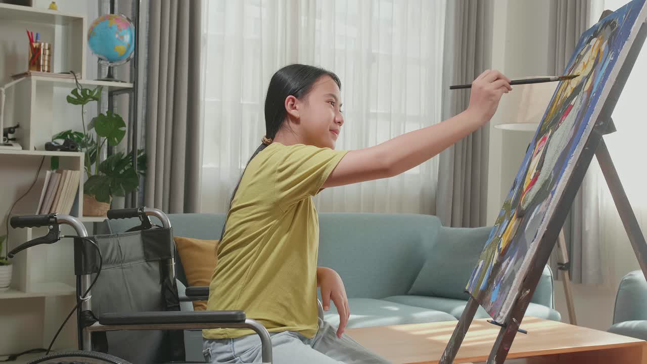 Side View Of An Asian Artist Girl In Wheelchair Holding Paintbrush And Painting A Girl On The Canvas