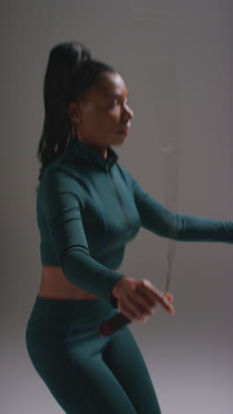 vertical video studio action shot of female athlete wearing tracksuit training in gym fitness class with jump rope or skipping rope against plain background