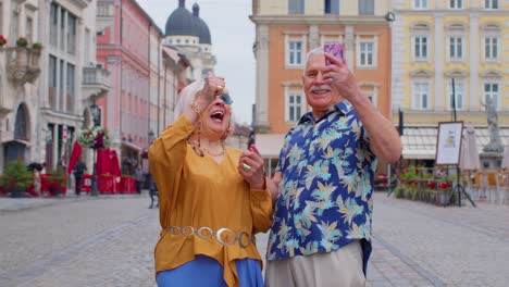 Turistas-Mayores-Y-Elegantes-Que-Buscan-Un-Teléfono-Inteligente-Sinceramente-Regocijándose-Por-Ganar,-Mensaje-Con-Buenas-Noticias