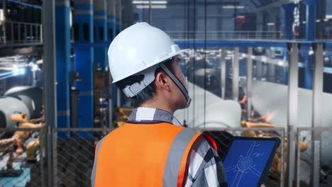 vista cercana de atrás de un ingeniero masculino asiático con casco de seguridad trabajando en una tableta con el plan de la turbina eólica en una fábrica de turbinas eólicas mientras el brazo robótico trabaja