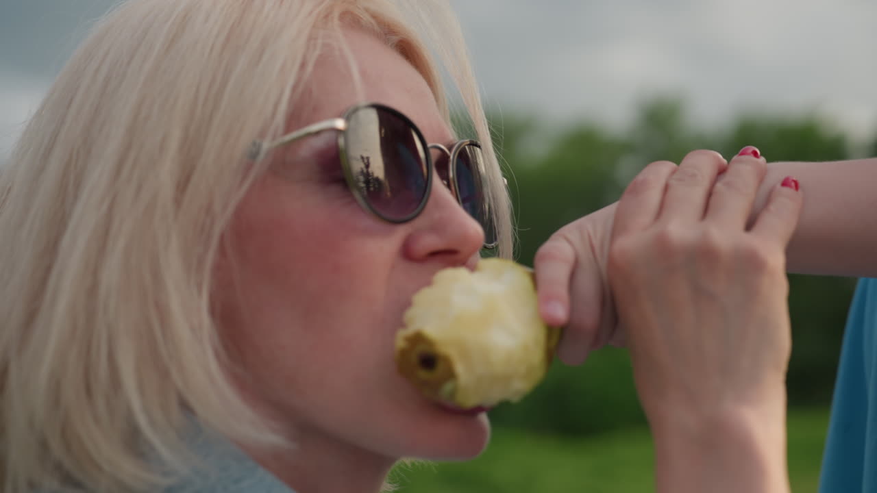 playful mom with red nails leaning back to eat pear offered by daughter, daughter holding fruit and eating snack, sunny outdoor picnic, loving playful gesture between mother and child in green park