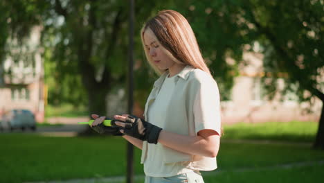 side view of young woman walking outdoors, examining green air pump with focused, thoughtful expression, sunlight reflects off her face, with blurred background of trees and distant building