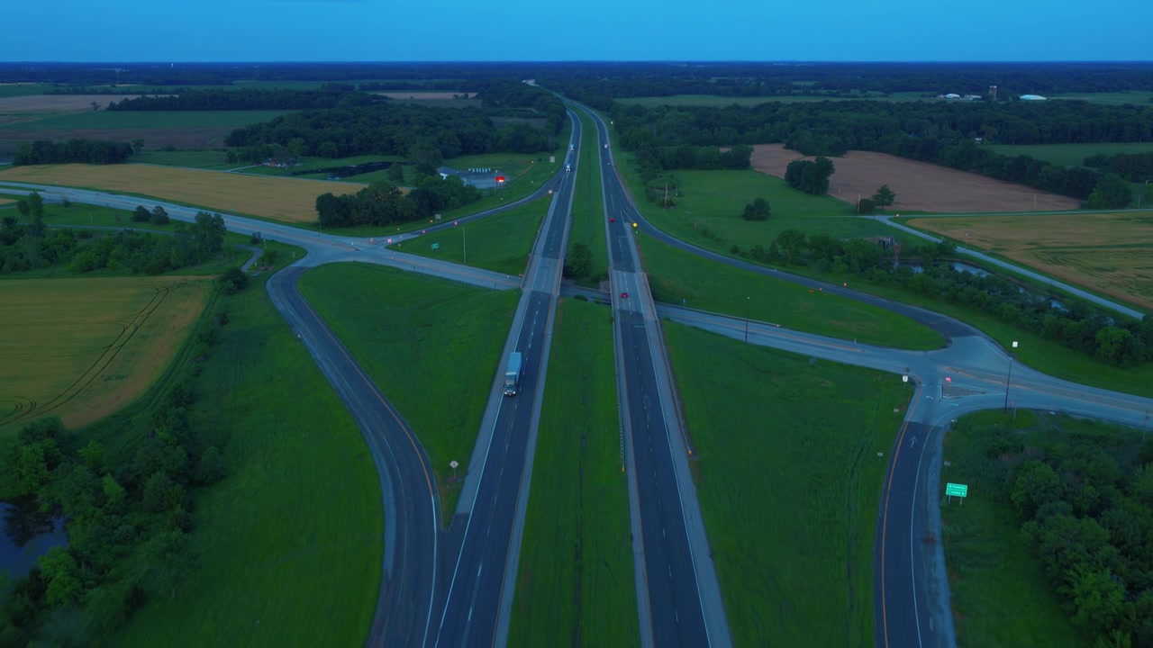 Drone tracking a semi truck on a divided highway at dusk, tractor trailer freight and traffic moving along the interstate during blue hour twilight beside a pond.