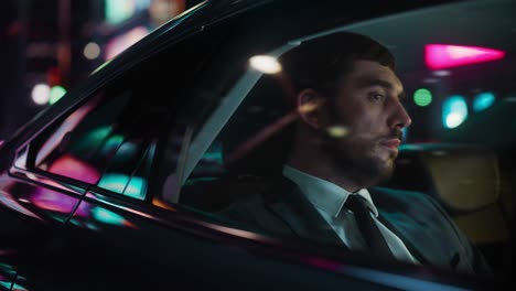 handsome businessman in a suit commuting from office in a backseat of his car at night. entrepreneur passenger traveling in a transfer taxi in urban city street with working neon signs.